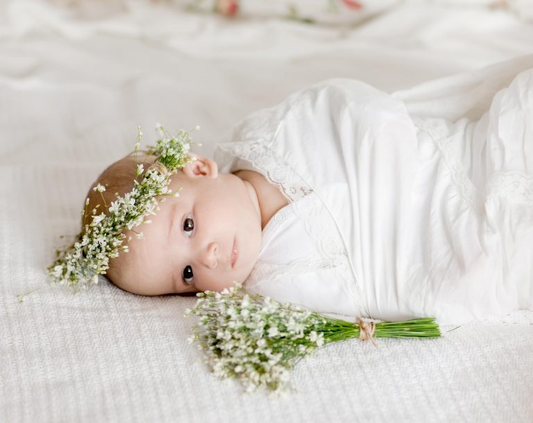 Baby swaddled in a white blanket, wearing a flower crown of baby's breath, laying on a bed