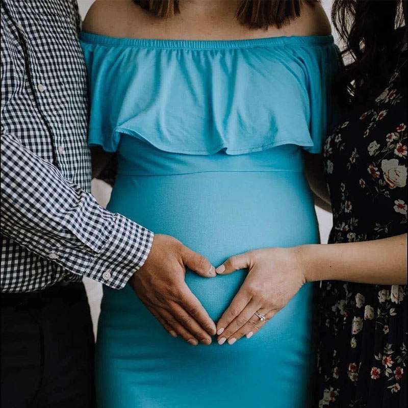 a surrogate with Canadian Fertility Consulting stands with her intended father and intended mother in a blue dress, with the intended parents place their hands on her growing pregnant belly making a heart shape