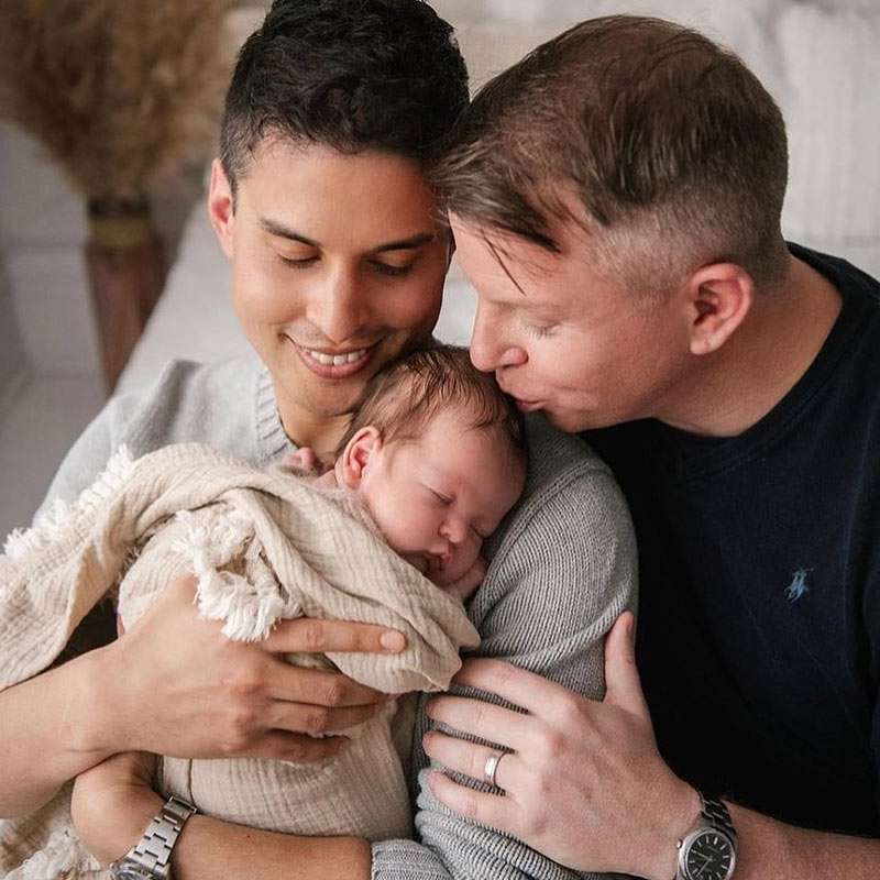 a newborn baby born via surrogacy in Canada sleeping over the shoulder of his dad with his other dad kisses his forhead