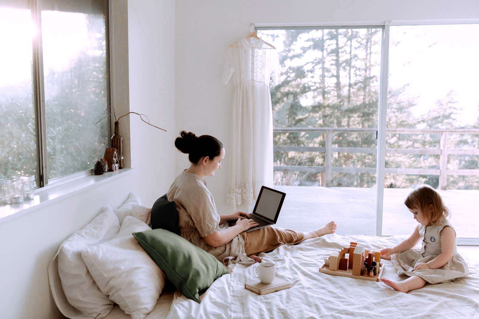 mother and 3 year old daughter on a cozy bed. Daughter is playing with blocks while the mother researches on her laptop with a cup of tea by her side.