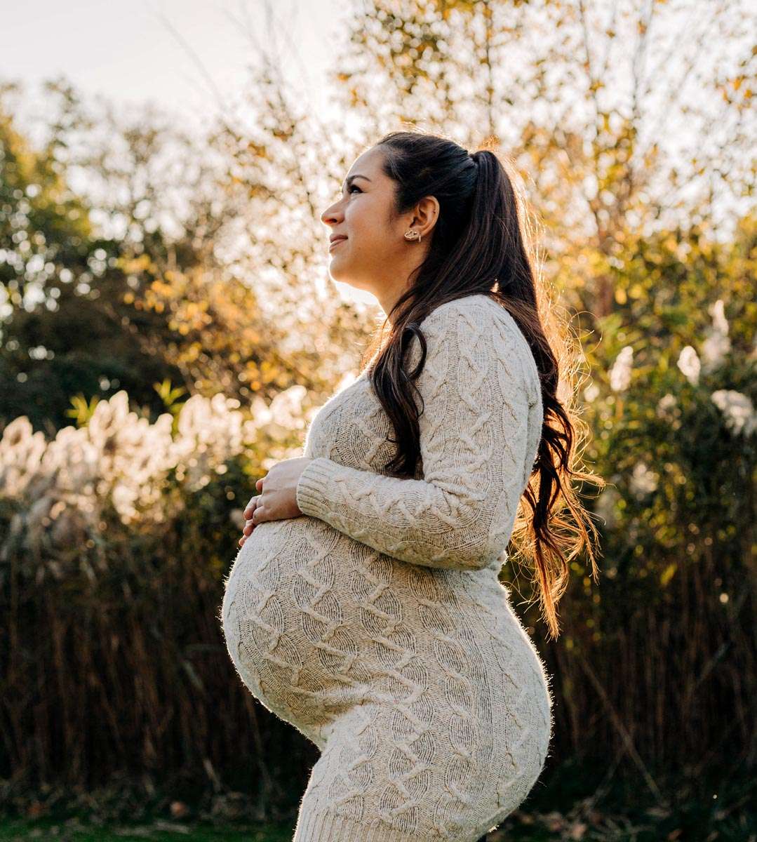 a pregnant surrogate with her hands on her growing baby belly smiling in front of a lush background of greenery