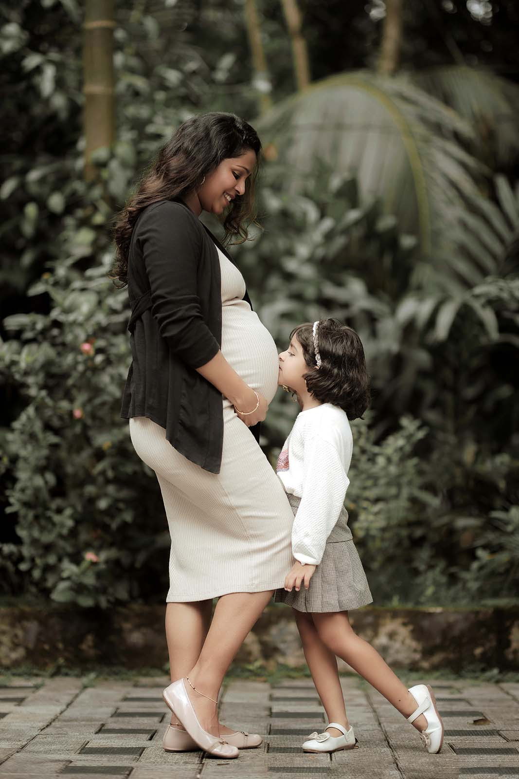 surrogate mother cradles her pregnant belly while her daughter leans in to kiss her mother’s growing baby bump. They are surrounded by luscious tropical plants and greenery.